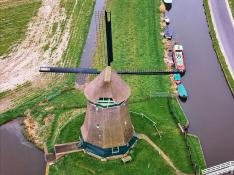 Aerial view of De Kaagmolen windmill with its thatched roof and sails situated beside a narrow canal with moored boats and green fields in Spanbroek North Holland Netherlands. Aerial view of De Kaagmolen windmill with its thatched roof and sails situated beside a narrow canal with moored boats and green fields in Spanbroek North Holland Netherlands.