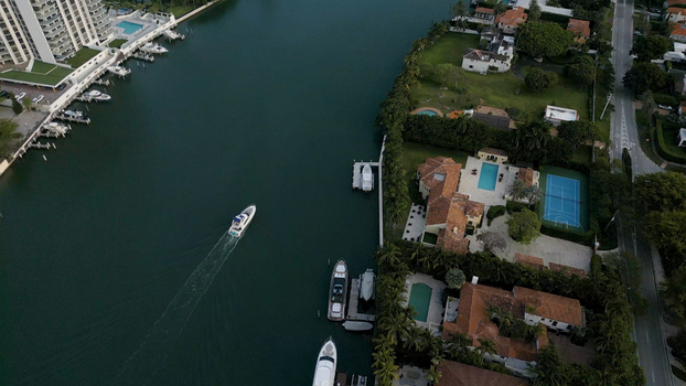 Aerial view of a motorboat cruising through a canal lined with luxury waterfront estates featuring swimming pools tennis courts and lush tropical landscaping in Miami Beach Florida United States. Aerial view of a motorboat cruising through a canal lined with luxury waterfront estates featuring swimming pools tennis courts and lush tropical landscaping in Miami Beach Florida United States.