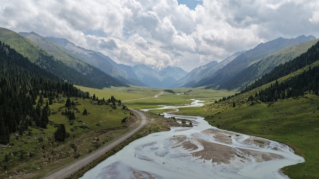 Aerial view of Enilchek valley featuring a winding river a dirt road lush green slopes pine forests and towering mountains under a cloudy sky in Enilchek Issyk Kul Region Kyrgyzstan. Aerial view of Enilchek valley featuring a winding river a dirt road lush green slopes pine forests and towering mountains under a cloudy sky in Enilchek Issyk Kul Region Kyrgyzstan.