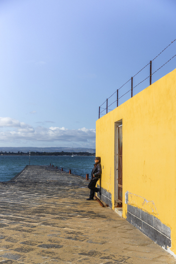 View of a lone figure leaning against a bright yellow wall next to a stone pier stretching into the tranquil blue sea under a clear sky Ortigia Sicily Italy.
