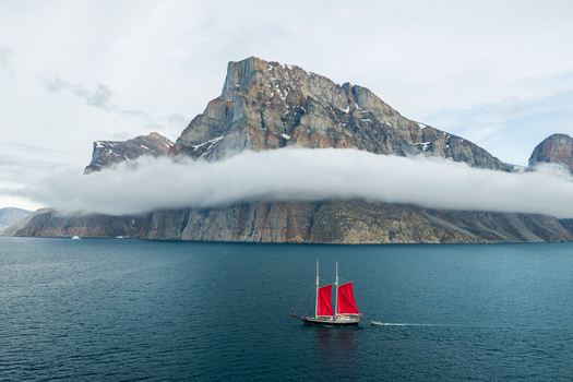 Aerial view of a sailboat with red sails navigating the calm waters of a fjord set against the towering Storoen mountain partially shrouded in clouds in Avannaata Kommunia Greenland. Aerial view of a sailboat with red sails navigating the calm waters of a fjord set against the towering Storoen mountain partially shrouded in clouds in Avannaata Kommunia Greenland.
