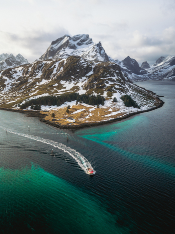 Aerial view of a boat slicing through the turquoise waters towards the snow dusted islands and rugged peaks of Kåkernbrua Ramberg Nordland Norway. Aerial view of a boat slicing through the turquoise waters towards the snow dusted islands and rugged peaks of Kåkernbrua Ramberg Nordland Norway.