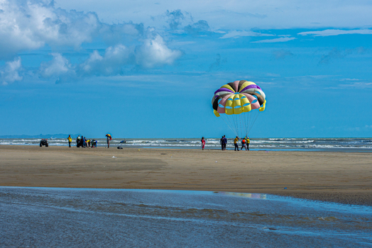 Cox s Bazar Bangladesh 18 August 2023 Aerial view of a colorful parasail drifting gently above the sandy beach as people gather at the shoreline where the blue sky meets the ocean. Cox s Bazar Bangladesh 18 August 2023 Aerial view of a colorful parasail drifting gently above the sandy beach as people gather at the shoreline where the blue sky meets the ocean.