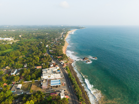 Aerial view of the tropical coastline featuring a coastal road lush palm trees and turquoise ocean waves crashing against the shore in Galle Southern Province Sri Lanka.