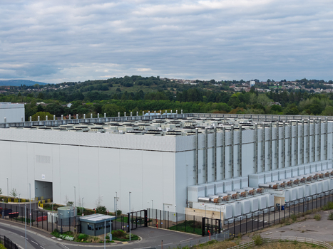 Aerial view of a large data center facility with cooling units on the roof security fencing and surrounding greenery in the United Kingdom.