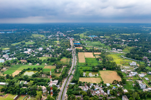 Aerial view of the city of Bogura featuring a main road through lush green fields buildings and communication towers under a cloudy sky in Bogura Rajshahi Division Bangladesh. Aerial view of the city of Bogura featuring a main road through lush green fields buildings and communication towers under a cloudy sky in Bogura Rajshahi Division Bangladesh.