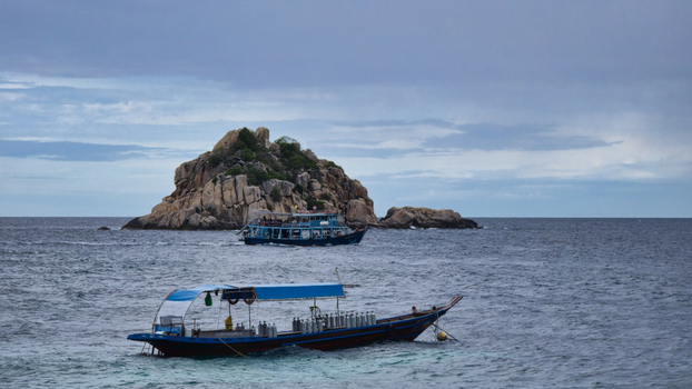 View of a traditional wooden boat equipped with scuba tanks floating on the sea with a rocky island in the background under a cloudy sky in Ko Tao Surat Thani Thailand.