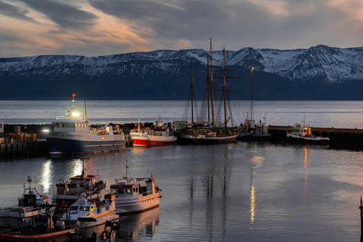 View of boats nestled in the harbor reflecting the twilight sky with snow capped mountains painting the horizon in hues of blue and gold Húsavík Norðurþing Iceland. View of boats nestled in the harbor reflecting the twilight sky with snow capped mountains painting the horizon in hues of blue and gold Húsavík Norðurþing Iceland.