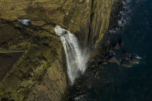 Aerial view of Mealt Falls cascading over the basalt cliffs of Kilt Rock into the dark blue sea with a faint rainbow in the mist Isle of Skye Scotland United Kingdom. Aerial view of Mealt Falls cascading over the basalt cliffs of Kilt Rock into the dark blue sea with a faint rainbow in the mist Isle of Skye Scotland United Kingdom.