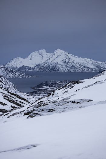 View of snow covered mountain peaks overlooking a calm fjord under a moody winter sky Kvaloya Troms Norway.