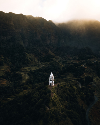 Aerial view of Capelinha de Nossa Senhora de Fatima perched on a lush green mountain ridge surrounded by terraced hills and misty slopes in the golden hour light in Sao Vicente Madeira Portugal.