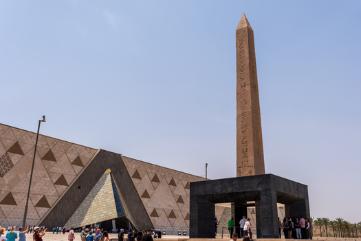 Giza Egypt 16 May 2026 View of the Grand Egyptian Museum featuring its modern triangular facade and a towering ancient obelisk with tourists gathered in the courtyard.