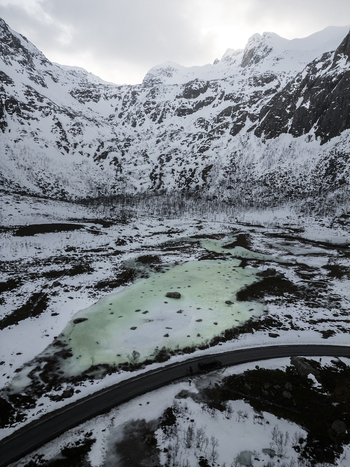 Aerial view of the snow covered mountains of Grotfjord and a frozen turquoise lake next to a winding road in Tromvik Troms Norway. Aerial view of the snow covered mountains of Grotfjord and a frozen turquoise lake next to a winding road in Tromvik Troms Norway.