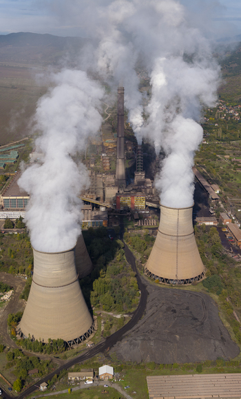Aerial view of a large coal fired power plant with cooling towers emitting thick white smoke into the sky surrounded by industrial infrastructure in Bulgaria.