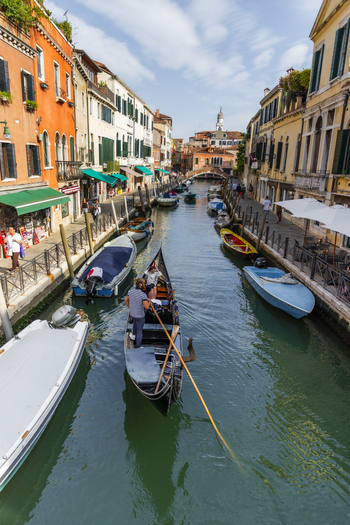 Venice Italy 21 September 2026 View of a gondolier rowing a gondola through a narrow canal surrounded by historic colorful buildings moored boats and pedestrians on stone pathways. Venice Italy 21 September 2026 View of a gondolier rowing a gondola through a narrow canal surrounded by historic colorful buildings moored boats and pedestrians on stone pathways.