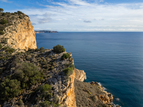 Aerial view of Cala Llebeig featuring rugged limestone cliffs Mediterranean Sea and sparse coastal vegetation under a blue sky in El Poble Nou de Benitatxell Valencian Community Spain.