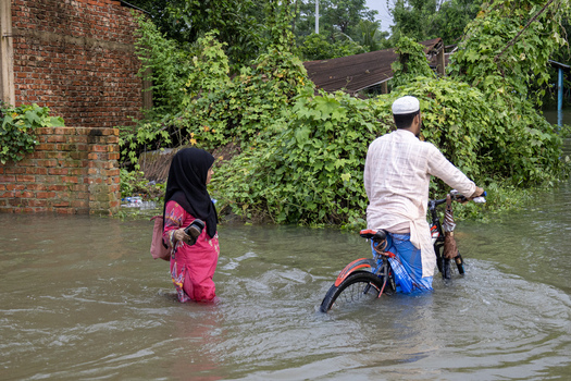 Banshkhali Upazila Chittagong Division Bangladesh 10 July 2026 View of a man on a bicycle and a woman walking through floodwaters in a rural area with brick walls and vegetation. Banshkhali Upazila Chittagong Division Bangladesh 10 July 2026 View of a man on a bicycle and a woman walking through floodwaters in a rural area with brick walls and vegetation.