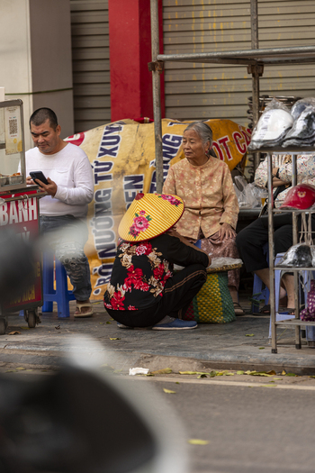 Hanoi Ha Noi Vietnam 11 November 2025 View of people sitting on plastic stools on a sidewalk with a woman in a conical hat and a man using a smartphone.