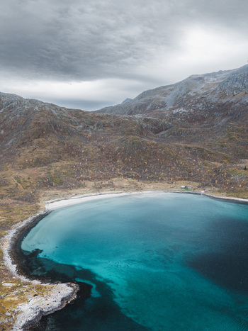 Aerial view of the turquoise waters of a bay surrounded by rocky autumn colored hills under a cloudy sky in Senja Troms Norway.
