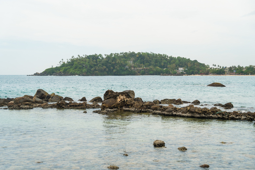 View of the rocky coastline and clear turquoise waters with a lush green tropical island in the background under a cloudy sky in Mirissa Southern Province Sri Lanka. View of the rocky coastline and clear turquoise waters with a lush green tropical island in the background under a cloudy sky in Mirissa Southern Province Sri Lanka.