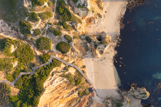 Aerial view of the rugged coastline and golden sandy beach of Lagos featuring dramatic limestone cliffs a wooden boardwalk and clear blue ocean waters in Lagos Faro District Portugal.