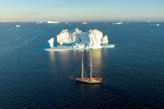 Aerial view of a large arch iceberg floating in the deep blue ocean with a classic sailing ship passing by under clear skies in Greenland. Aerial view of a large arch iceberg floating in the deep blue ocean with a classic sailing ship passing by under clear skies in Greenland.