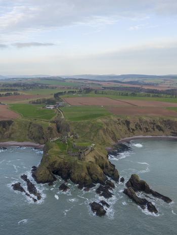 Aerial view of Dunnottar Castle perched on a dramatic rocky headland overlooking the North Sea with rolling green fields in the background Stonehaven Scotland United Kingdom. Aerial view of Dunnottar Castle perched on a dramatic rocky headland overlooking the North Sea with rolling green fields in the background Stonehaven Scotland United Kingdom.
