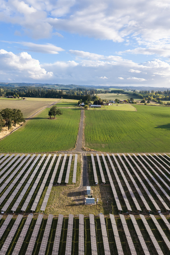 Aerial view of a large solar farm with rows of photovoltaic panels situated in a rural landscape with green fields trees and a blue sky in Woodburn Oregon United States. Aerial view of a large solar farm with rows of photovoltaic panels situated in a rural landscape with green fields trees and a blue sky in Woodburn Oregon United States.