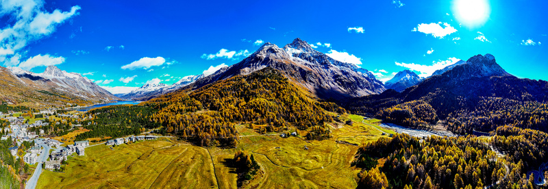 Aerial view of the Maloja District featuring golden autumn larch forests snow capped mountains a serene alpine lake and green meadows in Maloja District Grisons Switzerland.