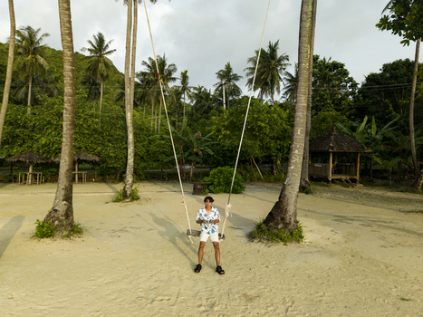Aerial view of a person enjoying a swing tied to palm trees on a sandy beach framed by lush greenery and distant huts Karimunjawa Jawa Tengah Indonesia.