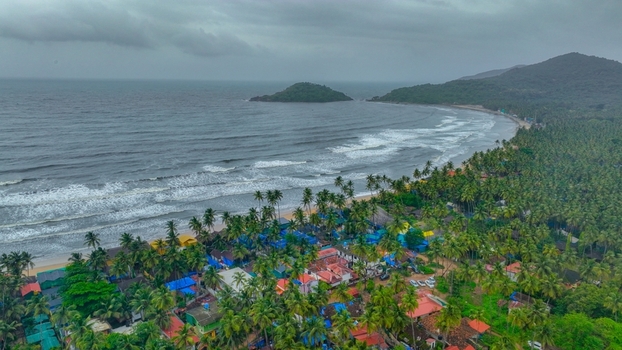 Aerial view of Palolem Beach with lush palm trees coastal village structures and ocean waves under a cloudy sky in Canacona Goa India.