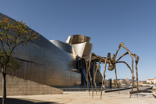View of the Guggenheim Museum Bilbao with its shimmering titanium panels and the iconic Maman spider sculpture under a clear blue sky in Bilbao Euskadi Spain. View of the Guggenheim Museum Bilbao with its shimmering titanium panels and the iconic Maman spider sculpture under a clear blue sky in Bilbao Euskadi Spain.