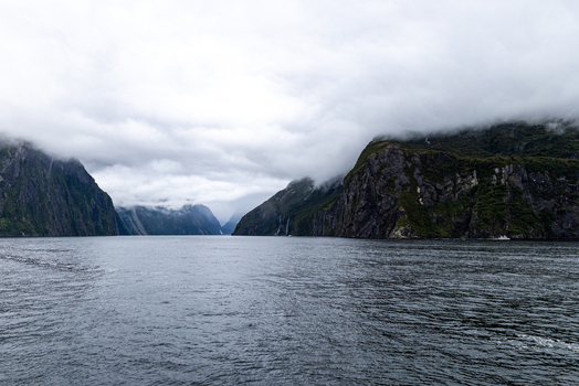 View of imposing mist shrouded cliffs rise dramatically from the still dark waters a symphony of gray and green under a vast cloudy sky Milford Sound Southland Region New Zealand.