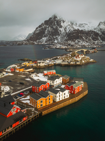 Aerial view of red and yellow cottages stand vibrant against the cold dark waters with snow capped mountains looming in the distance Henningsv&aelig;r Nordland Norway.