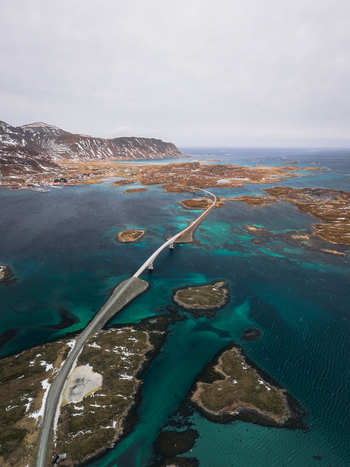 Aerial view of a long bridge cutting through the turquoise waters amidst islands creating a stark contrast with the snow capped mountains Flakstadbruene Nordland Norway. Aerial view of a long bridge cutting through the turquoise waters amidst islands creating a stark contrast with the snow capped mountains Flakstadbruene Nordland Norway.
