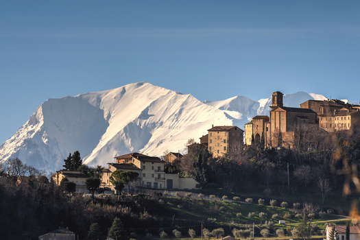 View of the historic town of Mogliano with stone buildings nestled against a backdrop of snow capped mountains under a clear blue sky in Mogliano Marche Italy.
