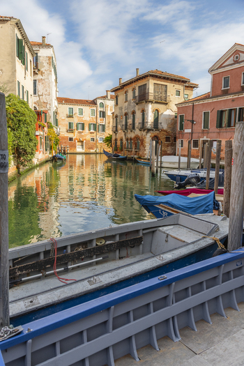 View of a quiet canal with traditional wooden boats moored along weathered historic buildings under a soft blue sky in Venice Veneto Italy. View of a quiet canal with traditional wooden boats moored along weathered historic buildings under a soft blue sky in Venice Veneto Italy.