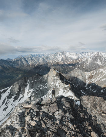 Aerial view of a person standing on a rocky mountain peak overlooking the vast snow capped peaks of the Alaska Wilderness in Palmer Alaska United States. Aerial view of a person standing on a rocky mountain peak overlooking the vast snow capped peaks of the Alaska Wilderness in Palmer Alaska United States.