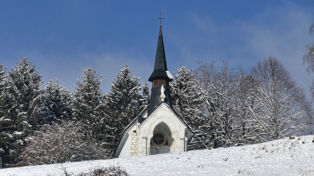 Aerial view of the small white chapel on a snow covered hill surrounded by frost covered trees under a clear blue sky in Vesancy Auvergne Rhone Alpes France. Aerial view of the small white chapel on a snow covered hill surrounded by frost covered trees under a clear blue sky in Vesancy Auvergne Rhone Alpes France.