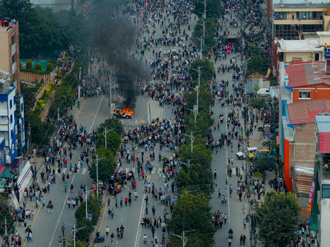Aerial view of a large crowd of people gathered on a city street with a vehicle engulfed in flames and thick black smoke rising surrounded by urban buildings and trees in Nepal.