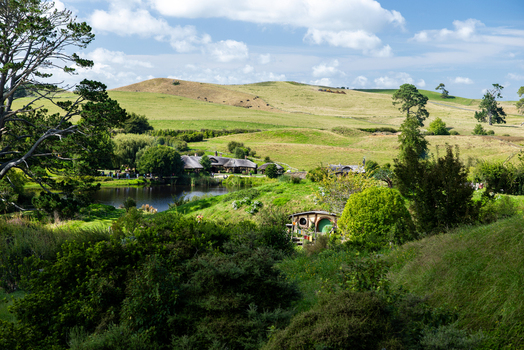 View of Hobbit holes nestled in rolling green hills a tranquil pond reflecting the sky framed by lush foliage Hobbiton Movie Set Waikato Region New Zealand. View of Hobbit holes nestled in rolling green hills a tranquil pond reflecting the sky framed by lush foliage Hobbiton Movie Set Waikato Region New Zealand.