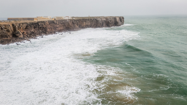 Aerial view of the historic Sagres Fortress perched on rugged cliffs with powerful waves crashing against the shore in Sagres Faro Portugal. Aerial view of the historic Sagres Fortress perched on rugged cliffs with powerful waves crashing against the shore in Sagres Faro Portugal.