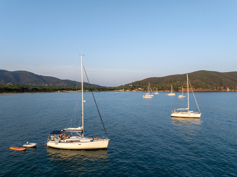 Aerial view of sailboats anchored in the calm blue waters of Lacona Bay surrounded by green hills under a clear sky in Lacona Tuscany Italy.