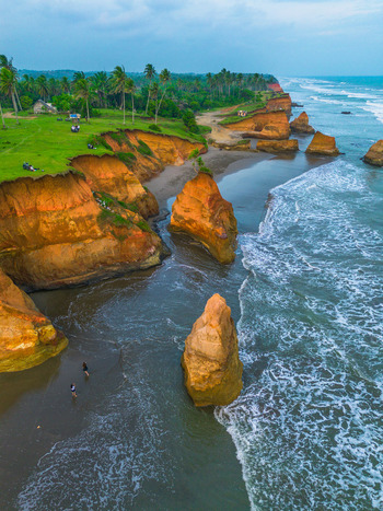 Aerial view of dramatic orange cliffs and lush green grassy coastline with palm trees crashing sea waves and people walking on the sandy beach in Bali Bali Indonesia. Aerial view of dramatic orange cliffs and lush green grassy coastline with palm trees crashing sea waves and people walking on the sandy beach in Bali Bali Indonesia.
