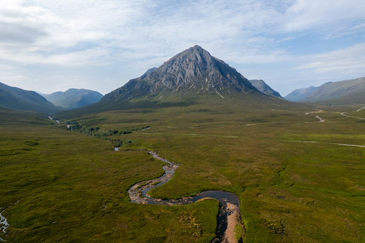 Aerial view of Buachaille Etive Mor mountain peak rising above the vast green landscape of Glen Etive with a winding river flowing through the valley in Ballachulish Scotland United Kingdom. Aerial view of Buachaille Etive Mor mountain peak rising above the vast green landscape of Glen Etive with a winding river flowing through the valley in Ballachulish Scotland United Kingdom.