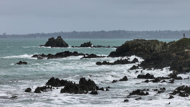Aerial view of the rugged coastline featuring dark jagged rocks and crashing white waves with a person standing on the cliff edge in Locquirec Brittany France.