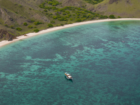 Aerial view of a wooden boat anchored in the turquoise waters of Komodo National Park with a sandy beach and rolling green hills in Komodo East Nusa Tenggara Indonesia. Aerial view of a wooden boat anchored in the turquoise waters of Komodo National Park with a sandy beach and rolling green hills in Komodo East Nusa Tenggara Indonesia.