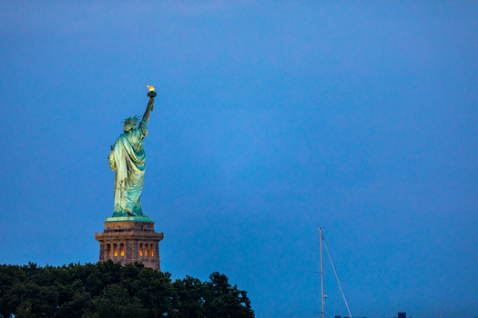 New York New York United States 08 July 2021 View of the Statue of Liberty illuminated against a deep blue twilight sky with trees at the base and a sailboat mast. New York New York United States 08 July 2021 View of the Statue of Liberty illuminated against a deep blue twilight sky with trees at the base and a sailboat mast.
