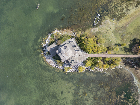 Aerial view of a building on a small peninsula in Lake Shkodra with a wooden pier surrounding trees and small boats in the clear water in Shiroka Shkoder County Albania. Aerial view of a building on a small peninsula in Lake Shkodra with a wooden pier surrounding trees and small boats in the clear water in Shiroka Shkoder County Albania.