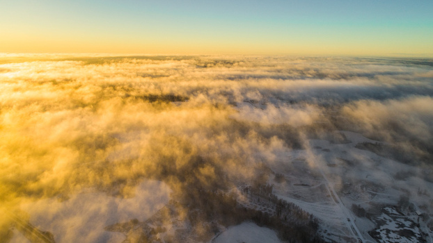 Aerial view of a landscape blanketed in thick morning mist during golden hour with snow covered fields and patches of forest visible below in Latvia. Aerial view of a landscape blanketed in thick morning mist during golden hour with snow covered fields and patches of forest visible below in Latvia.